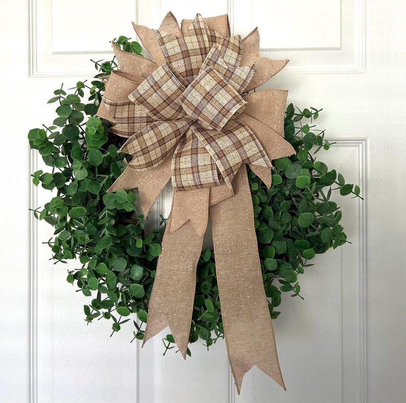 May include: A circular wreath composed of dense green eucalyptus leaves, featuring a large decorative bow at the top. The bow is crafted from a combination of solid tan burlap ribbon and a tan, brown, and white plaid patterned ribbon with long tails.