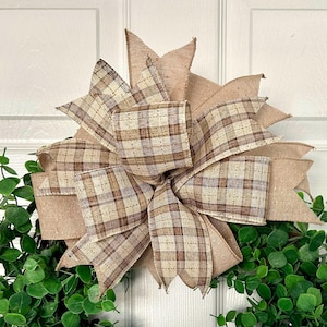 May include: A decorative bow with a plaid pattern in shades of beige and brown, attached to a wreath with green foliage. The bow is made of burlap and plaid ribbon, creating a rustic and elegant look. Perfect for seasonal home decor.