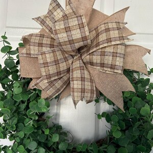 May include: A decorative wreath featuring a dense arrangement of small, rounded green leaves. A large, multi-layered bow is attached to the top, crafted from a combination of solid tan burlap ribbon and a brown and tan plaid patterned ribbon.
