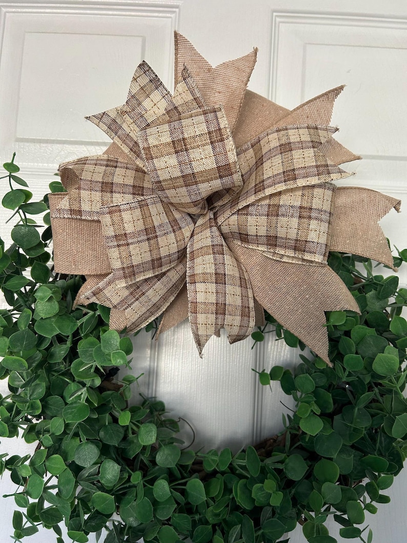 May include: A decorative wreath featuring a dense arrangement of small, rounded green leaves. A large, multi-layered bow is attached to the top, crafted from a combination of solid tan burlap ribbon and a brown and tan plaid patterned ribbon.