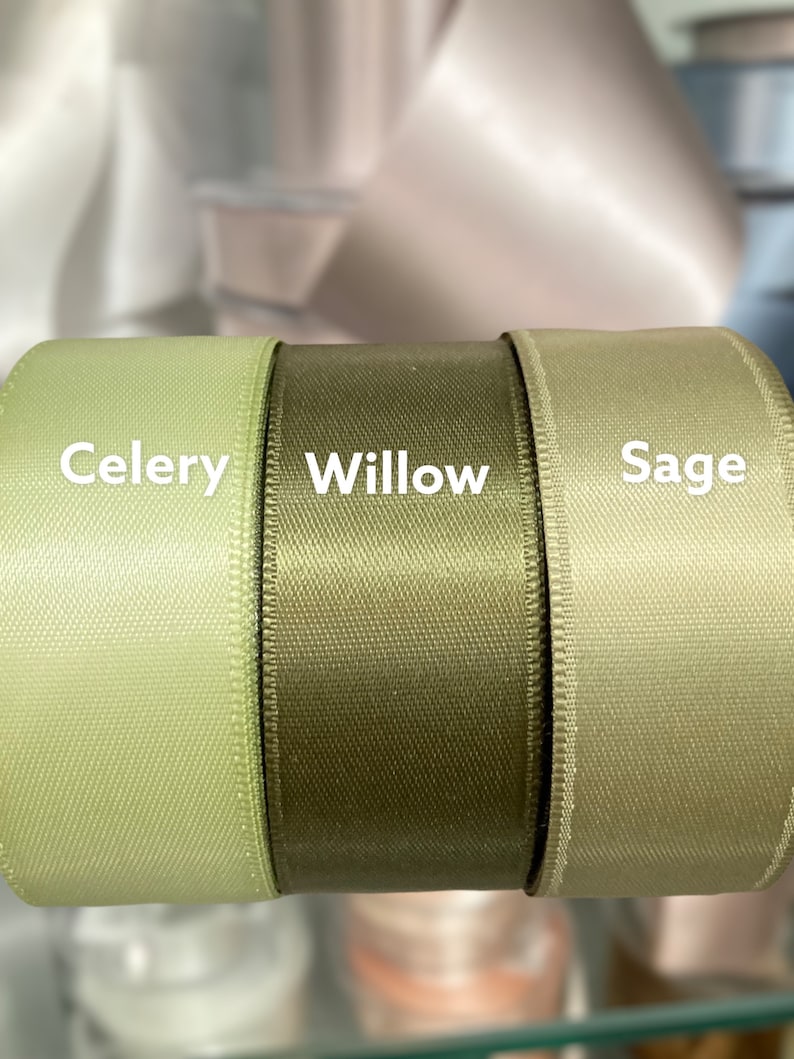 May include: Close-up of three spools of satin ribbon in shades of green: Celery, Willow, and Sage. The ribbon has a subtle sheen and a textured appearance. The names of the colors are printed in white on the ribbon.