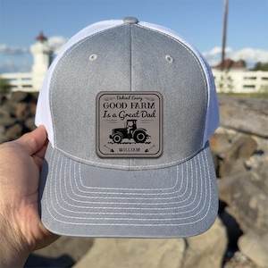 Op de afbeelding: Een grijze en witte truckerpet met een vierkante patch met een tractor en de tekst "Behind Every Good Farm Is a Great Dad". De pet heeft een grijze klep en kroon, met een witte mesh achterkant.