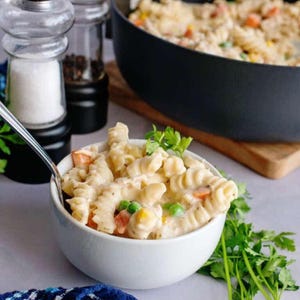 May include: A white bowl filled with creamy pasta, mixed with peas, carrots, and corn. A spoon rests in the bowl, with fresh parsley sprigs nearby. A pan of pasta and salt and pepper shakers are in the background.