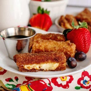 May include: A plate of French toast sticks, coated in cinnamon sugar, with a creamy filling. Served with a small metal cup of dark syrup, fresh strawberries, and blueberries. The food is on a white plate with a floral patterned cloth.