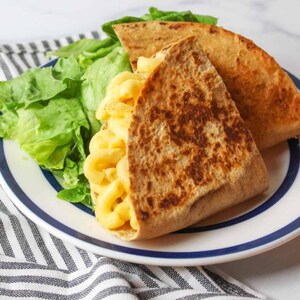 Puede incluir: Un plato con una quesadilla de macarrones con queso y una guarnición de lechuga. La quesadilla está hecha con una tortilla de trigo integral y rellena de macarrones con queso cremosos y amarillos. El plato tiene un borde a rayas azules y blancas.