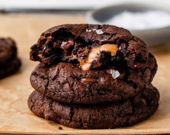 May include: A stack of three dark chocolate cookies with visible chocolate chips and a molten caramel center. The top cookie is broken open, revealing the soft interior. Coarse sea salt crystals are sprinkled on the dark brown surfaces.