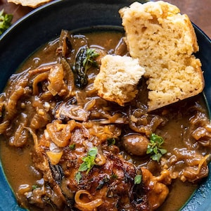 May include: A dark blue bowl containing a savory dish with caramelized onions, dark brown gravy, and a piece of cooked meat. Two pieces of light-colored, porous bread are placed on top of the stew, garnished with small green fresh herb leaves.