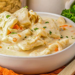 Puede incluir: Primer plano de un cuenco blanco lleno de pollo cremoso y albóndigas. Las albóndigas son grandes, de forma irregular, y el caldo tiene trozos visibles de zanahoria y perejil. El plato se sirve sobre una superficie de madera con un paño naranja.