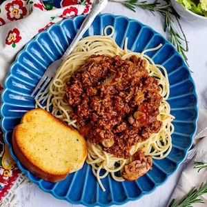 Könnte beinhalten: Spaghetti mit Fleischsauce und Pilzen, serviert mit Knoblauchbrot. Das Gericht befindet sich auf einem blauen Teller mit dekorativem Rand. Eine Gabel liegt auf dem Teller. Das Essen steht auf einer weißen Oberfläche mit einem floralen Tuch.