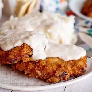 Op de afbeelding: Een bord met goudbruin gebakken kip, bedekt met romige witte jus. Zachte aardappelpuree vormt de bijgerecht. Een stevige maaltijd, een klassieker uit de Amerikaanse keuken.