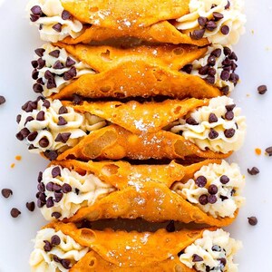 May include: Five golden-brown, tubular cannoli pastries arranged in a vertical row. Each pastry shell is filled with white, piped cream and studded with dark chocolate chips. A light dusting of powdered sugar covers the center of the pastries.