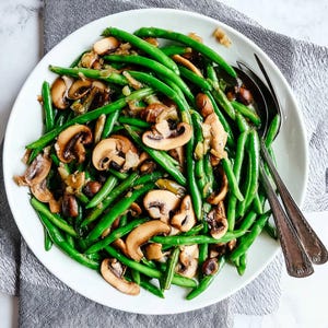 May include: A white plate filled with a vibrant green bean and mushroom dish. The green beans are long and slender, mixed with sliced brown mushrooms and herbs. Two silver serving utensils rest on the right side of the plate, with a gray cloth underneath.