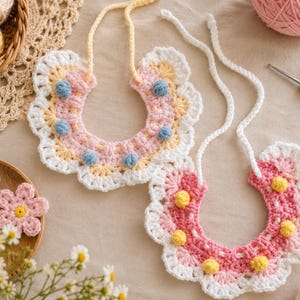 Pode incluir: Dois babadores de crochê feitos à mão com um design floral. Os babadores são rosa, branco, amarelo e azul. Os babadores têm uma borda ondulada e são decorados com pequenas bolas de crochê coloridas. Os babadores são exibidos com fio de crochê e flores.