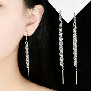 May include: Silver dangle earrings. Each earring has a series of interconnected, teardrop-shaped links that cascade downwards. A slender, straight bar hangs at the end of each earring, creating a sleek, modern look. The earrings are shown against a black background.