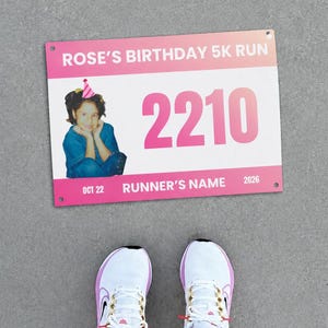 Pode incluir: Um número de peito retangular com design rosa e branco contendo o texto ROSE'S BIRTHDAY 5K RUN, o número 2210, a data OCT 22 e o texto RUNNER'S NAME 2026. Uma foto de uma criança com um chapéu de festa está no lado esquerdo.
