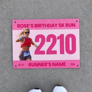 以下が含まれることがあります： ROSE'S BIRTHDAY 5K RUNの文字、大きなピンク色のフォントで書かれた2210の数字、ランナーの写真、RUNNER'S NAME、OCT 22、2026の文字が記載された長方形のピンクと白のレース用ゼッケン。下に白いスポーツシューズが見える。