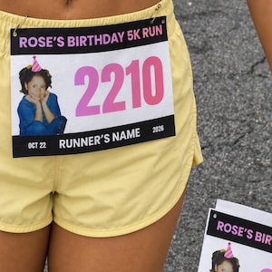Könnte beinhalten: Eine rechteckige Startnummer mit dem Text ROSE'S BIRTHDAY 5K RUN, der Nummer 2210 in großer rosa Schrift und einem Foto eines Kindes mit rosa Partyhut. Die Startnummer enthält den Text RUNNER'S NAME, OCT 22 und 2026 und ist an hellgelben Shorts befestigt.