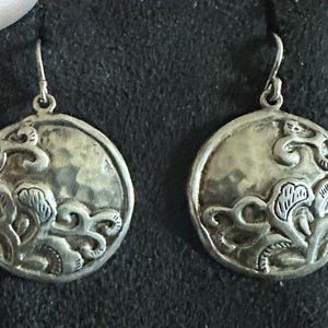 May include: A pair of round silver-toned drop earrings featuring a hammered texture background and raised, swirling organic patterns. Each circular earring is suspended from a simple silver-toned hook, showcasing a metallic finish with dark oxidation in the recessed areas of the intricate, embossed design.