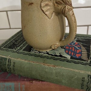 May include: A beige ceramic mug with a textured, speckled finish and a handle sculpted in the shape of an elephant head and trunk. The elephant design features carved details on the ears and forehead, with the trunk curving down to the base.