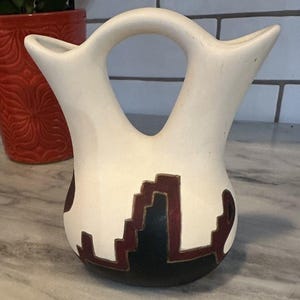 Puede incluir: Jarrón de cerámica de color crema con un diseño distintivo de doble pico y un asa central. El jarrón presenta un patrón geométrico en negro y burdeos, añadiendo un toque del suroeste. Una pieza decorativa para la decoración del hogar.