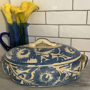 Könnte beinhalten: Eine ovale Auflaufform aus Keramik mit Deckel, die eine cremefarbene Basis und ein handgemaltes blaues Blumenmuster aufweist. Die Form hat zwei seitliche Griffe und einen zentralen Griff am Deckel sowie eine glänzende Oberfläche.