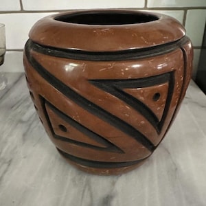 May include: A brown ceramic vase with a black geometric design. The vase has a wide, rounded shape with a slightly flared rim. The design features triangles and diagonal lines. The interior of the vase is black.
