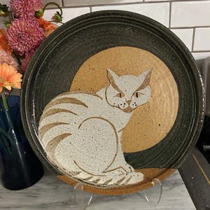 Pode incluir: Um prato redondo de cerâmica com um gato de cor esbranquiçada salpicada sentado de perfil contra um grande círculo de cor castanha sólida. O prato tem um rebordo texturado de cor cinzento-carvão e um acabamento salpicado no design.