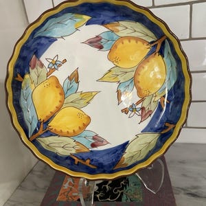 Puede incluir: Un plato decorativo de cerámica con borde festoneado. El plato presenta un diseño pintado a mano de limones amarillos, hojas verdes y azules y pequeñas flores sobre un fondo blanco. El borde es azul con un borde marrón y amarillo.