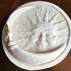 Pode incluir: Arte de parede circular branco sujo com um design de sol, lua e estrelas. O sol tem um rosto sorridente com raios e a lua tem formato de crescente. Três estrelas são visíveis no fundo. A peça de arte tem uma superfície texturizada.