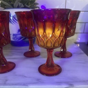 Puede incluir: Juego de cinco copas de vino tinto rubí vintage. Las copas tienen forma de tulipán con un patrón de diamantes en el cuenco y un tallo. El degradado de color pasa de un rojo intenso a un ámbar dorado. Las copas están sobre una superficie de mármol.