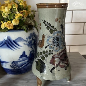 Puede incluir: Un jarrón de cerámica verde claro con borde marrón y tres pequeños pies. El jarrón está decorado con diseños florales azules, marrones y granates y un pequeño pájaro. En el fondo, una maceta de cerámica azul y blanca con flores amarillas.