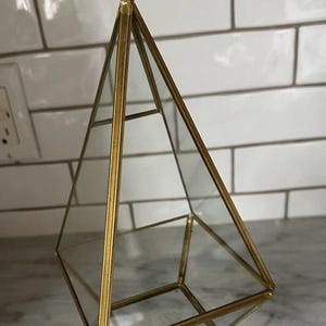 May include: A geometric terrarium with a pyramid shape, featuring clear glass panels and a gold-colored metal frame. The terrarium is suspended by a chain and ring, suitable for displaying small plants or decorative items. The design is modern and minimalist.
