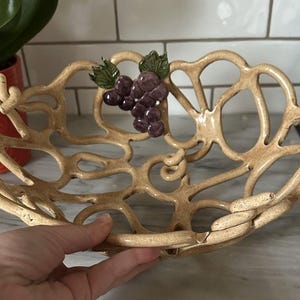 May include: Tan ceramic fruit bowl with an openwork design. The bowl features a cluster of purple grapes with green leaves. The bowl is held by a person's hand. The bowl is a decorative item.