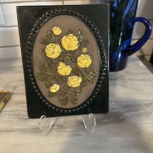 May include: A ceramic tile with a raised floral design. The tile has a dark green border and an oval centre. The centre displays yellow roses and green leaves against a brown background. The tile is supported by a clear stand.