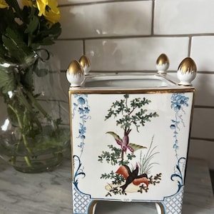 May include: A square, white ceramic planter with a gold rim and feet. The planter features a hand-painted design of birds and foliage in shades of green, red, and brown. The top corners have gold-tipped finials. A vase of yellow flowers is in the background.