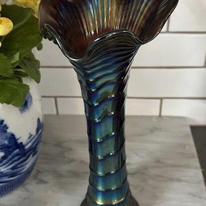 May include: An iridescent glass vase with a ruffled rim. The vase transitions from a deep burgundy at the top to shades of teal and gold down the ribbed body. The vase is a decorative item.