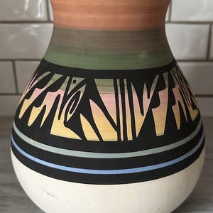 May include: A ceramic vase with a flared rim, featuring a horizontal color gradient of peach, green, and yellow at the top. The midsection displays a bold black band with abstract geometric cutouts, followed by thin blue and black stripes above a white base.