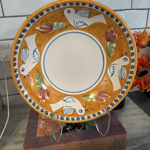 May include: A round ceramic bowl featuring a hand-painted design of four white birds interspersed with red and green floral motifs on an ochre background. The rim is decorated with a blue dotted pattern, and the center is a plain cream circle.
