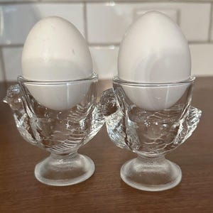May include: Two white eggs in clear glass egg cups. The egg cups are bird-shaped with detailed feather designs. They have a round base and a small bowl for the eggs. The background features a light-coloured tiled wall and a wooden surface.