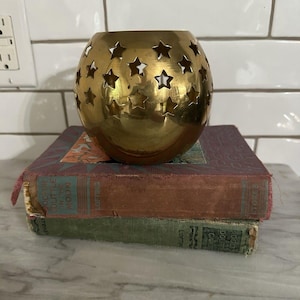 May include: A spherical gold-toned metal candle holder featuring numerous star-shaped cutouts across its surface. The object is resting on top of two stacked vintage hardcover books with worn edges and visible text on their spines.