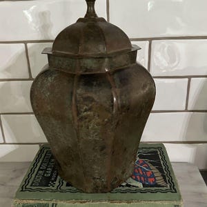 May include: A dark, weathered metal canister with a multi-faceted body and a matching lid topped with a finial. The canister rests on two stacked books, the top one titled Doctor Dolittle's Garden by Hugh Lofting, featuring a green cover with illustrations.