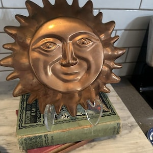 May include: A circular copper wall plaque featuring a raised, three-dimensional sun face with a smiling expression and radiating pointed rays. The metallic object has a warm, polished finish and includes a small metal loop at the top for hanging.