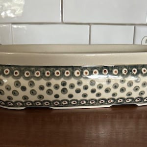 May include: A ceramic, oval-shaped planter with a cream-colored rim and a patterned exterior. The pattern features dark blue and gray polka dots with small red accents. The planter sits on a dark brown surface.