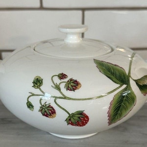 May include: White ceramic teapot with a lid and curved handle. The teapot is decorated with a strawberry vine design featuring red strawberries and green leaves. The teapot is on a marble surface.