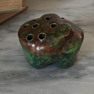 May include: A ceramic flower frog with a mottled green and brown glaze. The frog has a rounded shape with multiple holes on the top for arranging flowers. The base is a darker green.
