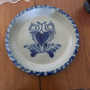Puede incluir: Un plato redondo de gres con un diseño azul y blanco. El plato presenta un diseño central de corazón con dos pájaros y un borde azul. El plato está sobre una superficie de madera.
