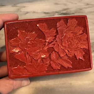 May include: A rectangular cinnabar red box featuring a detailed relief carving of two blooming peony flowers with leaves. The background of the lid displays a geometric lattice pattern, and the entire surface has a smooth, matte finish.