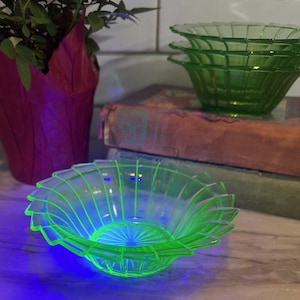 Può includere: Un set di quattro ciotole in vetro verde traslucido con un motivo a coste radiali e un bordo smerlato. Il vetro emette una brillante luce al neon, che indica la presenza di uranio, con una ciotola posizionata in primo piano e tre impilate.