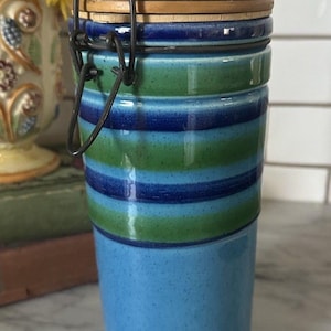 Puede incluir: Un tarro de cerámica con tapa de madera y cierre metálico. El cuerpo presenta rayas horizontales azules y verdes, con una base azul claro lisa. La tapa es de color marrón claro.
