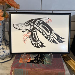 May include: A framed print featuring a stylized black and white eagle illustration with salmon-colored accents. The eagle is depicted in a traditional formline art style with bold black lines, curved shapes, and a sharp beak, set against a cream background.